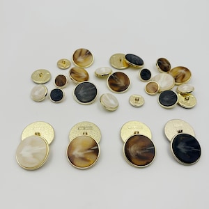 May include: Assortment of decorative buttons in various sizes and colors. The buttons feature gold rims and a range of colors, including cream, brown, black, and gold. Some buttons have a marbled appearance, while others are solid.
