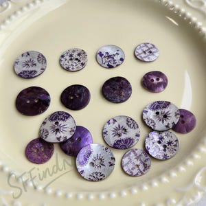 May include: A selection of decorative buttons in various sizes and designs. The buttons feature a mix of purple and white floral patterns, along with solid purple buttons. They are arranged on a light cream plate.