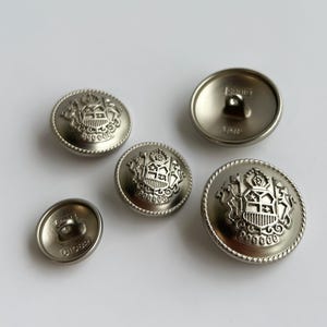 Puede incluir: Un conjunto de botones metálicos plateados de varios tamaños. Cada botón presenta un diseño de escudo en relieve detallado con una corona y un escudo. Los botones tienen un acabado metálico brillante, sobre un fondo blanco.