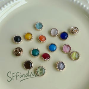 May include: A collection of colorful, round buttons with gold-colored metal rims. The buttons feature various colors, including blue, pink, yellow, and green. The image also includes the text "SFFindNZ" in a green, handwritten font.