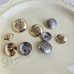 May include: A collection of decorative metal buttons in gold and silver tones. The buttons feature embossed crest and emblem designs. Various sizes and shapes are displayed on a light yellow surface. Some buttons have the text "28010" and "JDB" on the back.