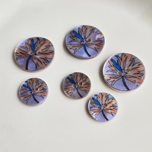 May include: Five round buttons of varying sizes, each featuring a light purple background with a printed design of a stylized tan and blue flower. Every button has two small holes in the center for attachment, arranged in a circular pattern.