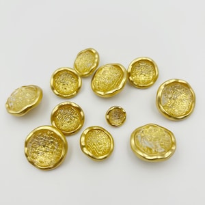 May include: A collection of gold-colored buttons with a textured, semi-transparent center and a gold rim. The buttons vary in size, with a small button in the center. The buttons are arranged on a white surface.