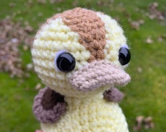 May include: A crocheted yellow duck with brown accents and black eyes. The duck is sitting in a hand.