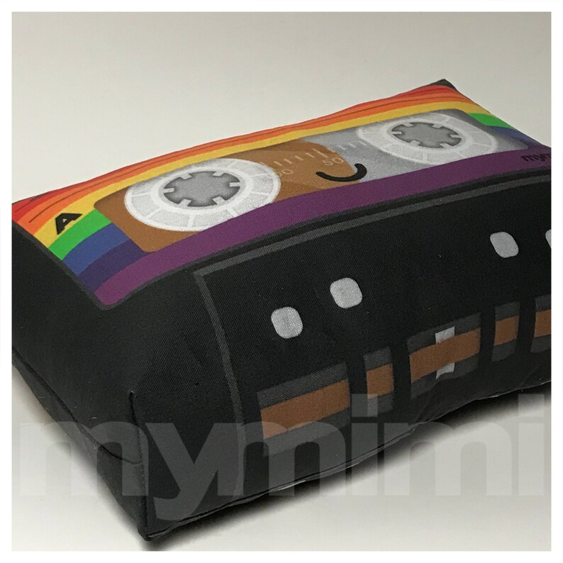 May include: A black and brown cassette tape pillow with a rainbow design on the top. The pillow has a smiling face and the letter "A" on the left side.