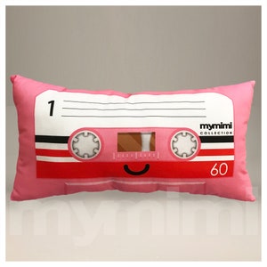 Op de afbeelding: Roze kussen in de vorm van een cassettebandje met de tekst "mymimi COLLECTION" en "60" erop gedrukt. De cassettebandje heeft een glimlachend gezicht.