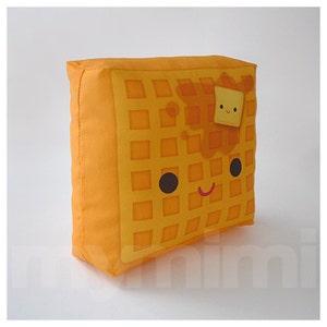 May include: A yellow waffle pillow with a smiling face, brown syrup, and a small yellow square of butter on top.