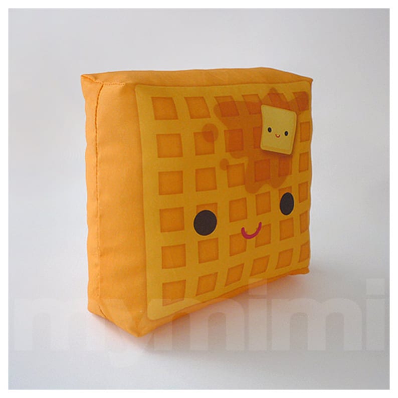 May include: A yellow waffle pillow with a smiling face, brown syrup, and a small yellow square of butter on top.