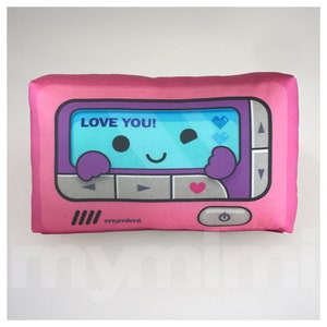 May include: A pink plush pillow shaped like a retro device, featuring a screen with a smiling face and the text "LOVE YOU!". The screen displays pixelated hearts. The pillow has a power button and the brand name "mymimi".