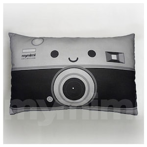 Op de afbeelding: Een grijs en zwart kussen met een cartoon camera-ontwerp. De camera heeft een glimlachend gezicht en de tekst "mymimi collection" is linksboven afgedrukt.