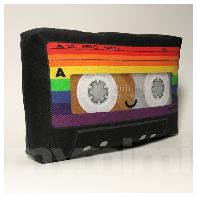 May include: A black cassette tape pillow with a rainbow colored design. The tape has a smiley face on it and the text "SIDE 1 STEREO NOISE RED." and "mymix".