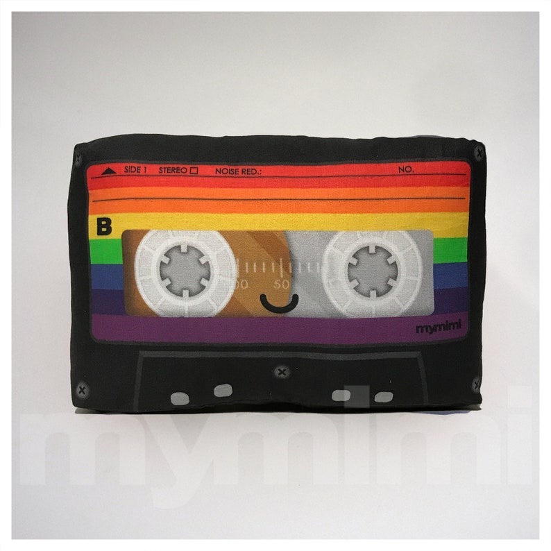 May include: A black cassette tape pillow with rainbow colored stripes and a smiling face. The pillow has the text "SIDE 1 STEREO NOISE RED.: NO. mymimi" printed on it.