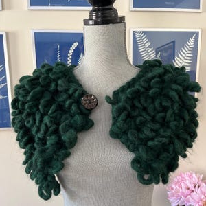 May include: A dark green, hand-knitted shrug with a textured, looped design. The shrug is fastened with a decorative, dark metal button. The garment is displayed on a mannequin, showcasing its unique, cozy style.