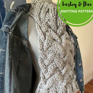 May include: A gray knitted cowl with a cable knit pattern. The cowl is draped over a denim jacket.