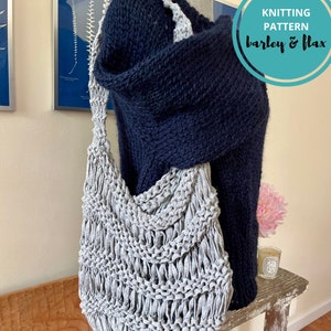 May include: A grey knitted tote bag with a wide, open top. The bag is made with a simple stitch pattern and has a relaxed, slouchy shape. The bag is hanging on a wooden surface, next to a dark blue knitted sweater.