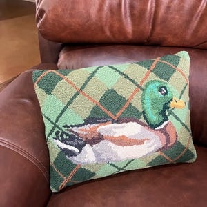 May include: A green and brown patterned cushion with a needlepoint design of a mallard duck. The cushion is sitting on a brown leather sofa.