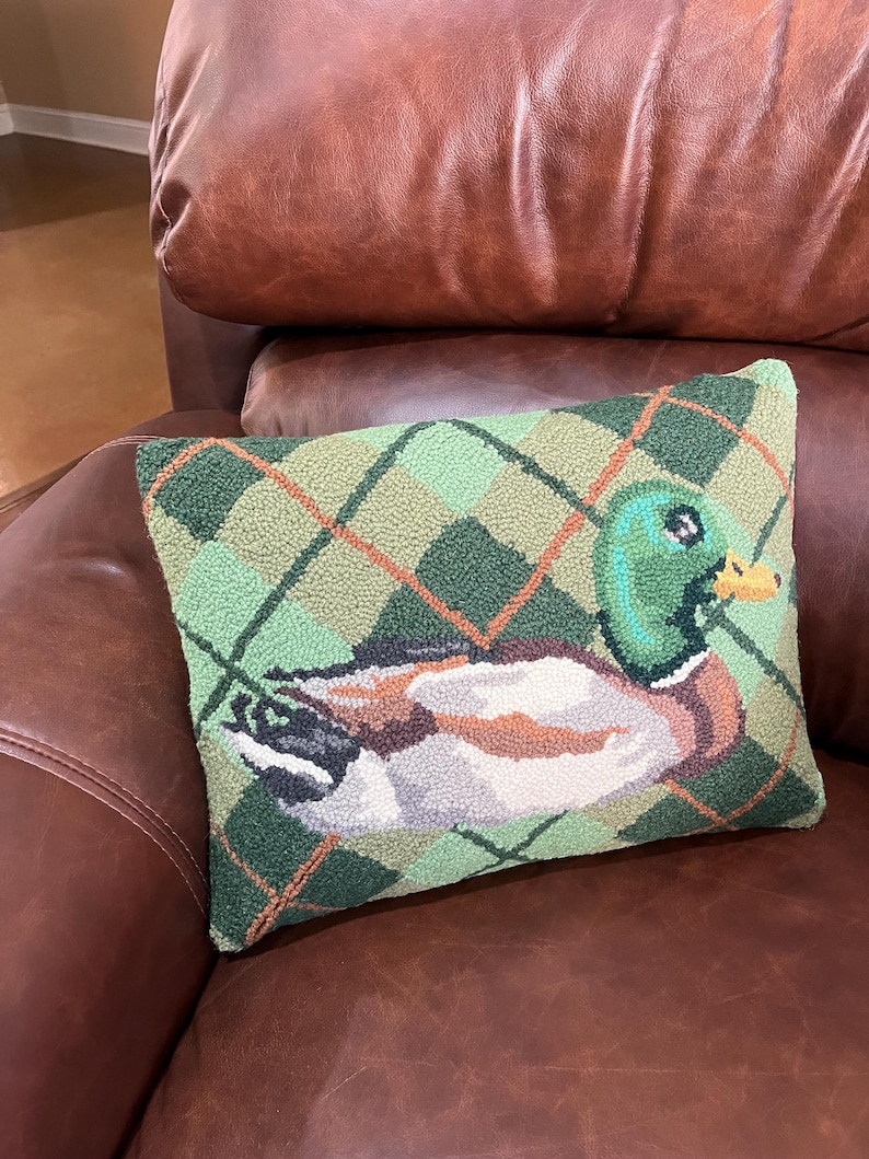 May include: A green and brown patterned cushion with a needlepoint design of a mallard duck. The cushion is sitting on a brown leather sofa.