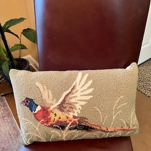 May include: A green throw pillow with a needlepoint design of a pheasant in flight. The pheasant is brown, orange, and blue with white wings.