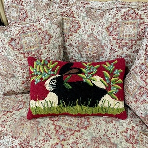 May include: A red cushion with a black and white rabbit design. The rabbit is surrounded by green leaves and white flowers. The cushion is on a sofa with a floral pattern.