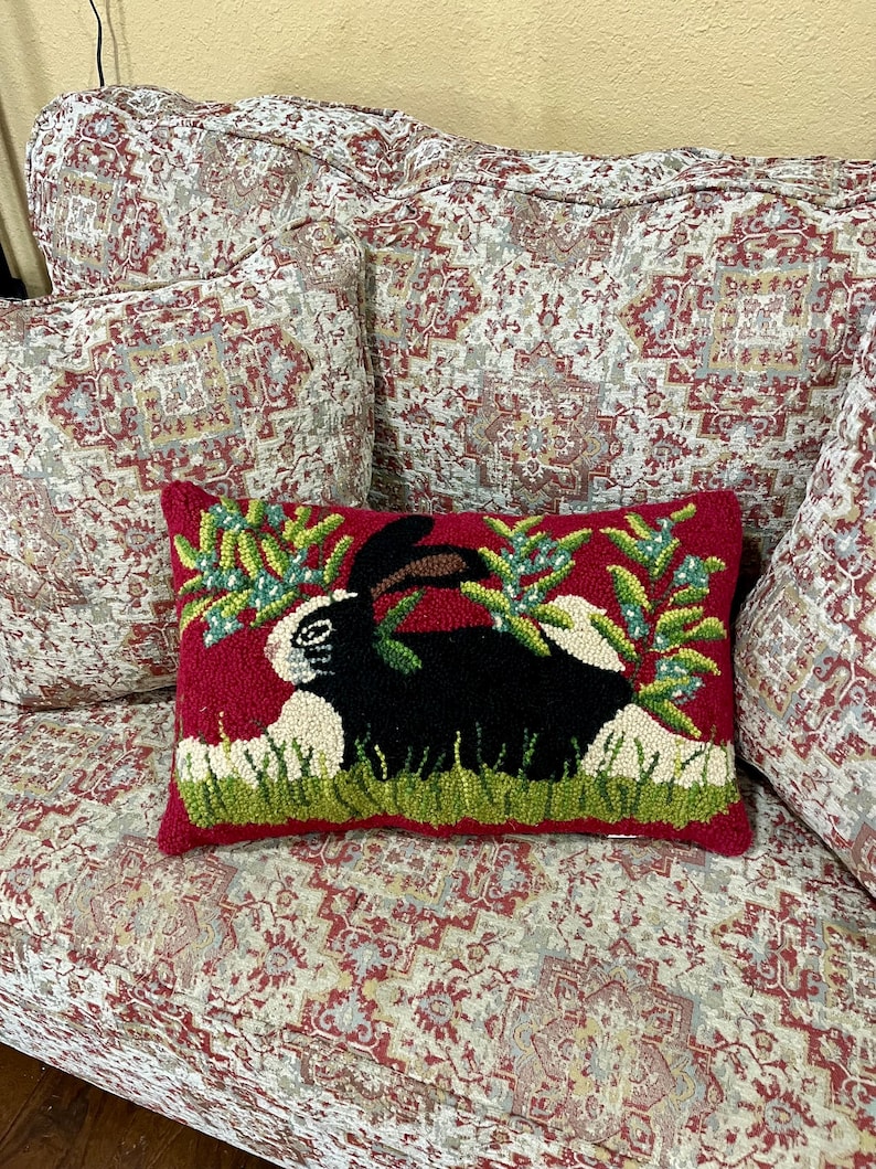 May include: A red cushion with a black and white rabbit design. The rabbit is surrounded by green leaves and white flowers. The cushion is on a sofa with a floral pattern.