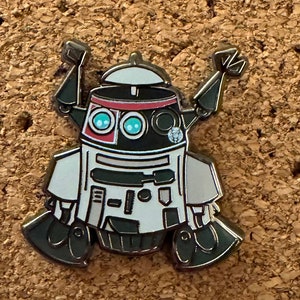 May include: A white, black, and red robot pin with a single black eye and two blue eyes. The robot has two arms that are raised in the air. The robot is standing on a brown corkboard.