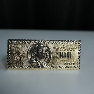 May include: A gold-colored rectangular object resembling a $100 bill. The design includes a portrait, the number "100", and intricate patterns. The object is likely a decorative item or accessory.