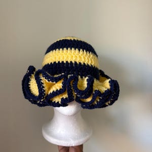 May include: A handmade crochet hat with a striped pattern in yellow and navy blue. The hat features a ruffled brim, adding a decorative element. The hat is displayed on a white mannequin head.