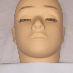 May include: A tan-colored mannequin head with closed eyes and attached eyelashes. The mannequin has light brown eyebrows and a smooth, neutral skin tone. The head is positioned above a white surface.