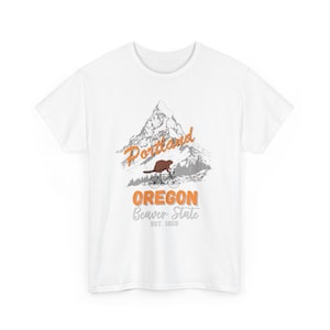 May include: White t-shirt with a graphic design of a mountain range with the text "Portland, Oregon, Beaver State, Est. 1859". A beaver is riding a bicycle in front of the mountain.