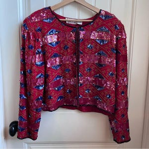 May include: A red sequined jacket with a geometric pattern of blue and pink sequins. The jacket has long sleeves and a round neckline. The edges are trimmed with black beading. The jacket is hanging on a wooden hanger.