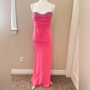 May include: A bright pink formal gown on a mannequin. The bodice features a sequined bust and thin spaghetti straps. The skirt is a long, flowing style. The dress is displayed in a well-lit room with a neutral background.