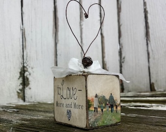 May include: A small wooden cube with a white background and the text "to love - more and more" printed on it. A small heart is printed below the text. A family of four is pictured walking in a field in the background. A wire heart with a brown bell is attached to the top of the cube.