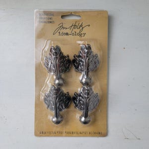 Puede incluir: Un paquete de cuatro patas metálicas oscuras para cajas de Tim Holtz idea-ology. El empaque es de cartón color canela con el texto Foundations, Fondations, Fundaciones y 4 Box Feet / 4 Pieds pour Boite / 4 Pies de Cuadro impreso.