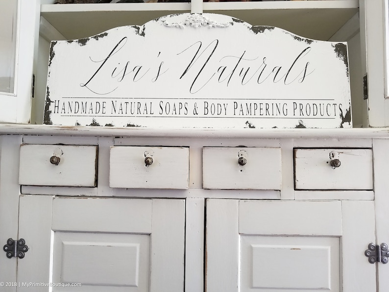 May include: White painted cabinet with four drawers and two doors. A sign above the cabinet reads "Lisa's Naturals Handmade Natural Soaps & Body Pampering Products."