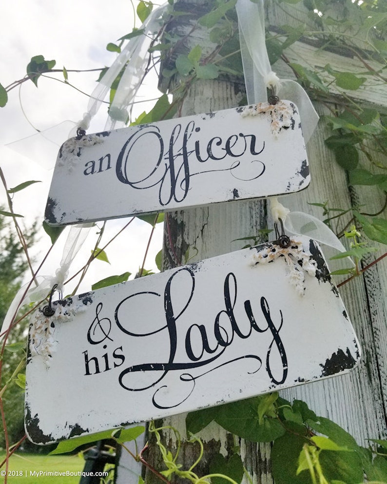Pode incluir: Duas placas retangulares brancas com letras pretas. A placa superior diz "an Officer" e a inferior "& his Lady". Decoradas com detalhes florais e fitas, ideais para um casamento ou evento especial.