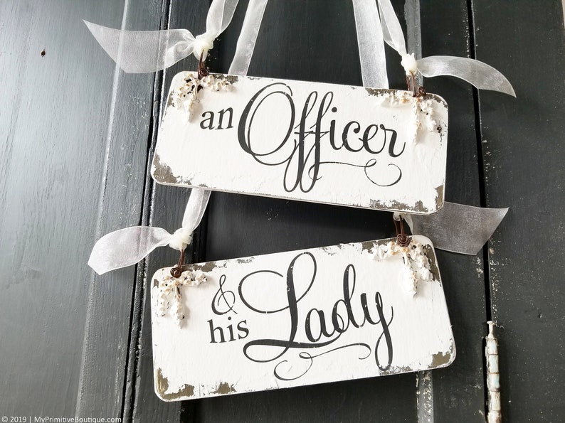 Pode incluir: Duas placas retangulares de madeira branca desgastada com o texto preto "an Officer" e "& his Lady". As placas s&atilde;o adornadas com pequenos detalhes florais e fita branca para pendurar.