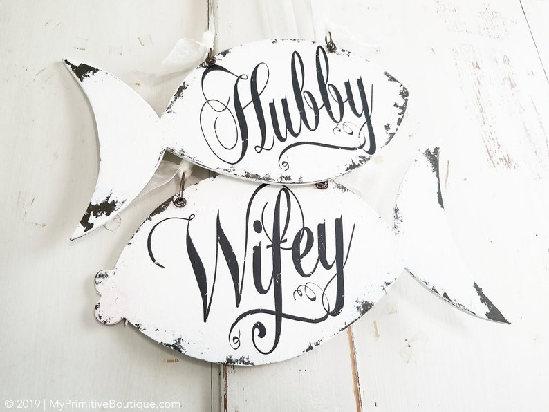 May include: Two white wooden fish-shaped signs with distressed paint. The top sign says "Hubby" and the bottom sign says "Wifey".