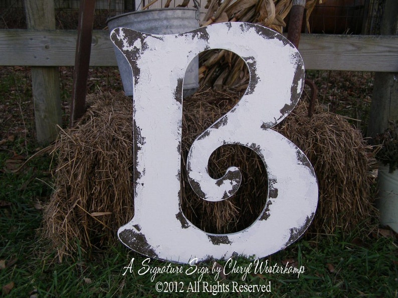 May include: A large, white, wooden letter B with a distressed, aged finish. The letter is standing on a pile of hay.