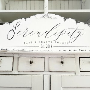 May include: White wooden sign with black lettering that reads "Serendipity Lash & Beauty Lounge Est. 2018".