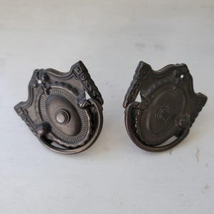 Peut inclure: Deux poignées de tiroir assorties en métal de couleur bronze foncé, dotées d'une plaque arrière ornée en forme de bouclier avec un médaillon central circulaire et une poignée tombante incurvée. La quincaillerie présente des détails en relief.