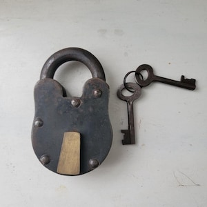 May include: A heavy, dark iron padlock with a rounded shackle and a central brass keyhole cover plate. The padlock features four visible rivets on its face. Two matching iron skeleton keys are attached to a small metal ring beside the lock.