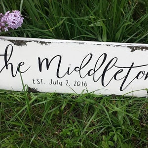 May include: White wooden sign with black painted text that reads "The Middletons Est. July 2, 2016". The sign is distressed and has a rustic look.