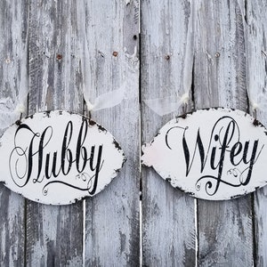 May include: Two white wooden fish-shaped signs with black lettering. The sign on the left says "Hubby" and the sign on the right says "Wifey".