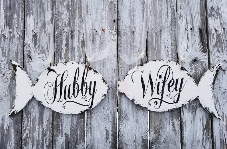 May include: Two white wooden fish-shaped signs with black lettering. The sign on the left says "Hubby" and the sign on the right says "Wifey".