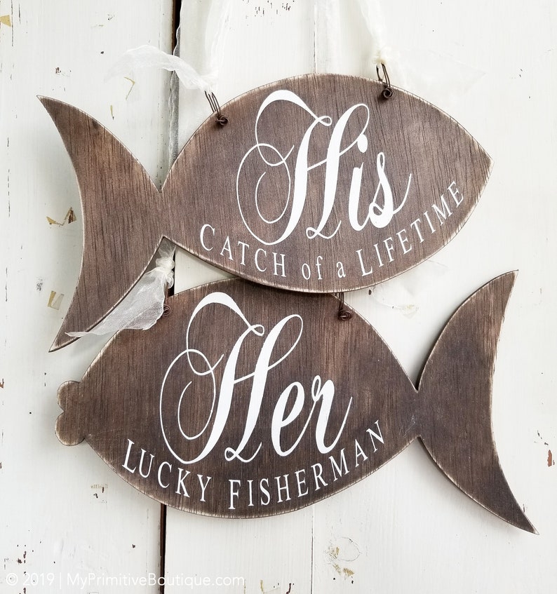 Puede incluir: Dos letreros de madera con forma de pez con texto blanco. El letrero superior dice "His Catch of a Lifetime". El letrero inferior dice "Her Lucky Fisherman".