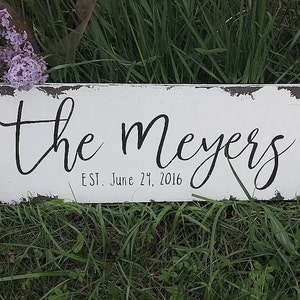 May include: White wooden sign with black lettering that reads "the meyers est. June 27, 2016". The sign has a distressed, rustic look and is sitting on a bed of green grass.