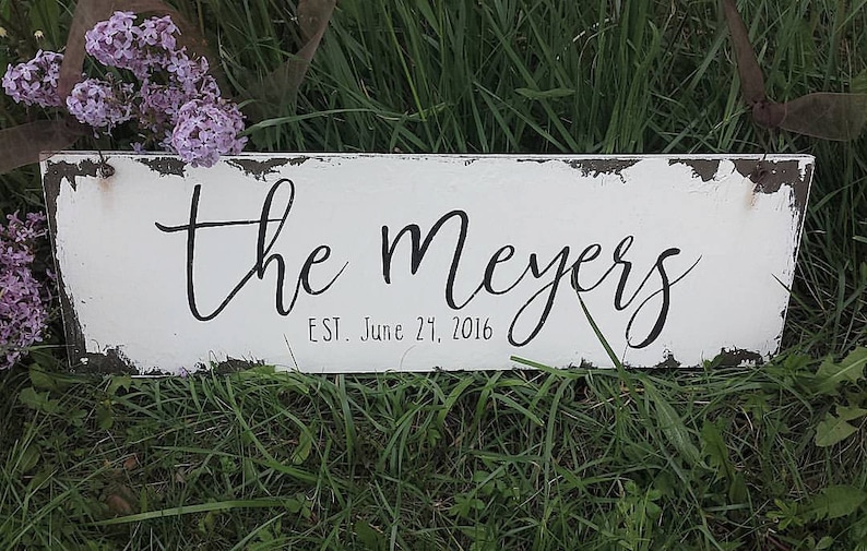 May include: White wooden sign with black lettering that reads "the meyers est. June 27, 2016". The sign has a distressed, rustic look and is sitting on a bed of green grass.