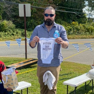 May include: A man wearing sunglasses and a blue shirt is holding a white onesie with the text "Future Carpenter Union Ironworker Local 11" printed on it.