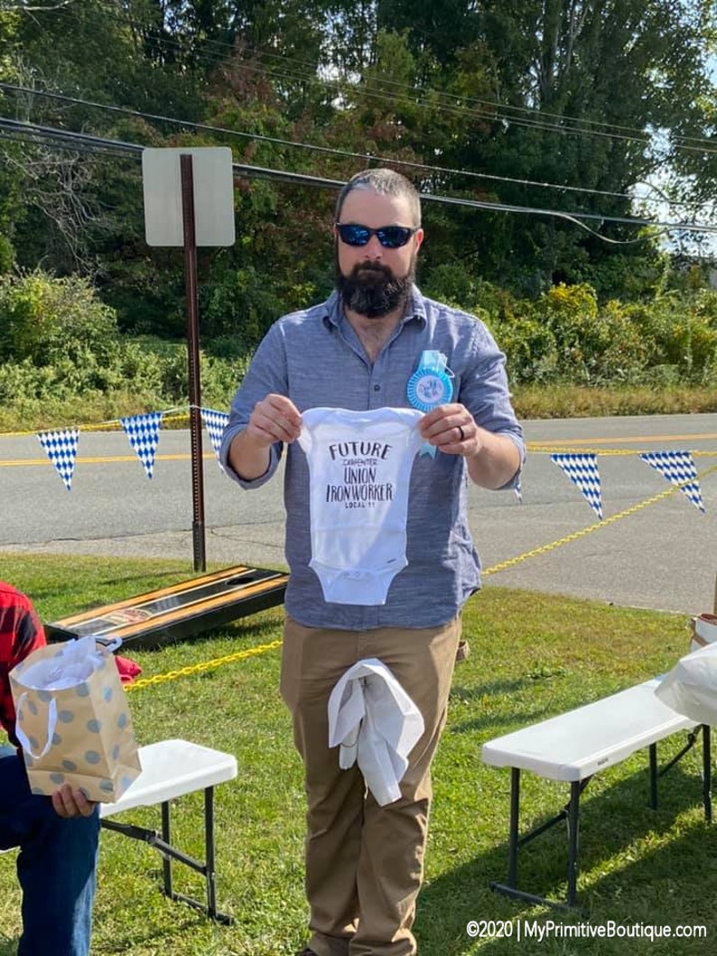 May include: A man wearing sunglasses and a blue shirt is holding a white onesie with the text "Future Carpenter Union Ironworker Local 11" printed on it.