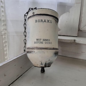 May include: A vintage off-white metal Boraxo powder dispenser with a cylindrical body, a black metal chain attached to the top, and a small black knob at the bottom. The text BORAXO and WET HANDS BEFORE USING are printed in black on the front surface.